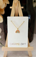 Gold necklace with a rainbow pendant on a Caramelsky card, displayed on a small wooden easel.