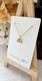 Gold necklace with a rainbow pendant on a Caramelsky branded card, displayed on a small wooden stand.
