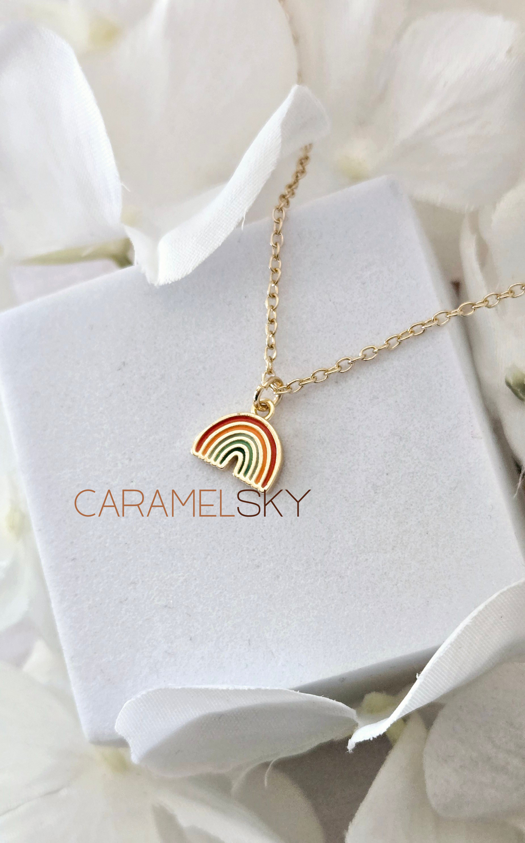 Gold necklace with a rainbow pendant on a white stand with 'Caramelsky' branding, surrounded by white flowers.