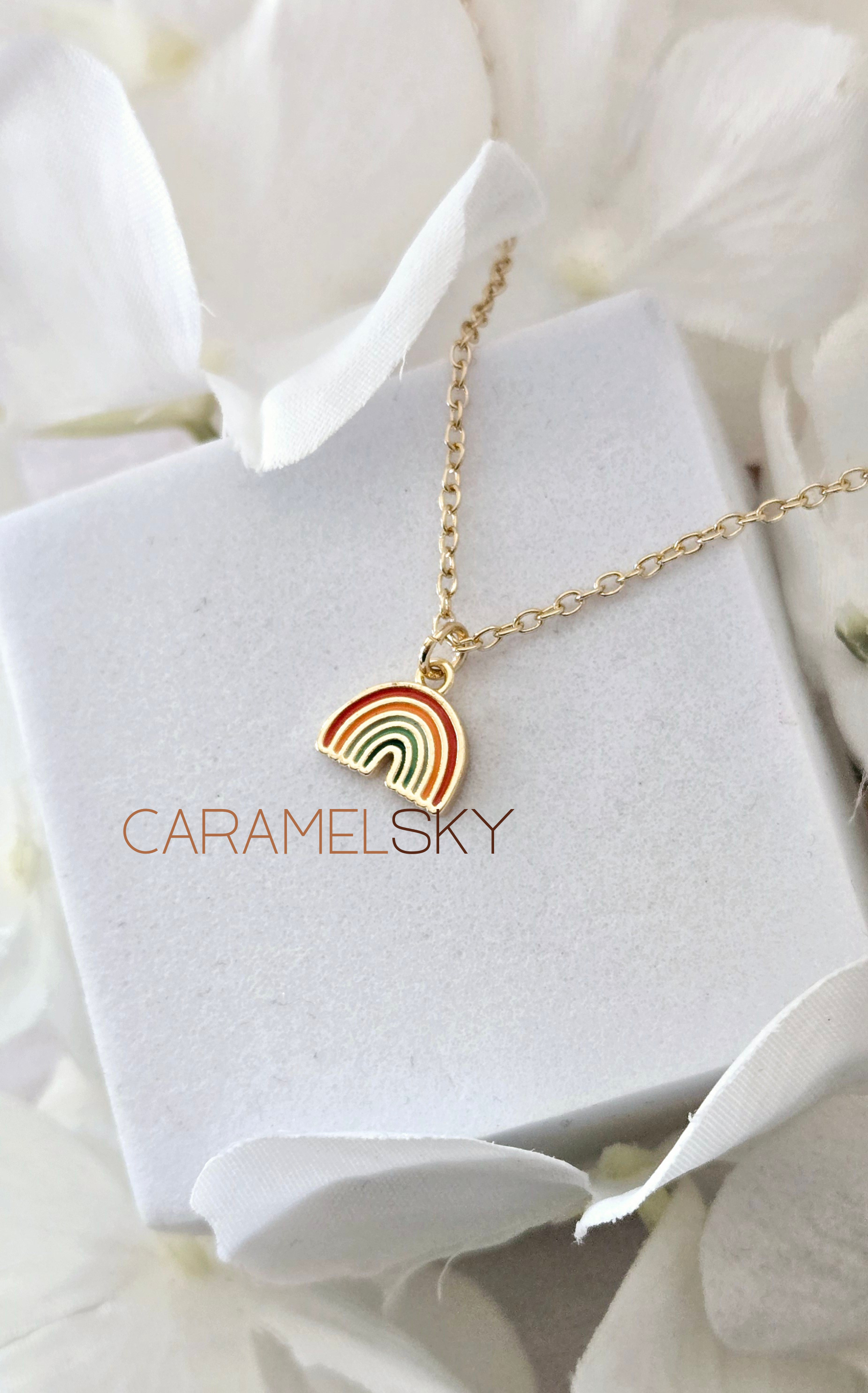 Gold necklace with a rainbow pendant on a white stand with 'Caramelsky' branding, surrounded by white flowers.